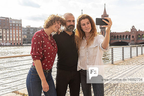 Mann und Freundinnen beim Selbstfahren mit dem Smartphone auf Brücke  Fluss und Gebäuden im Hintergrund  Berlin  Deutschland