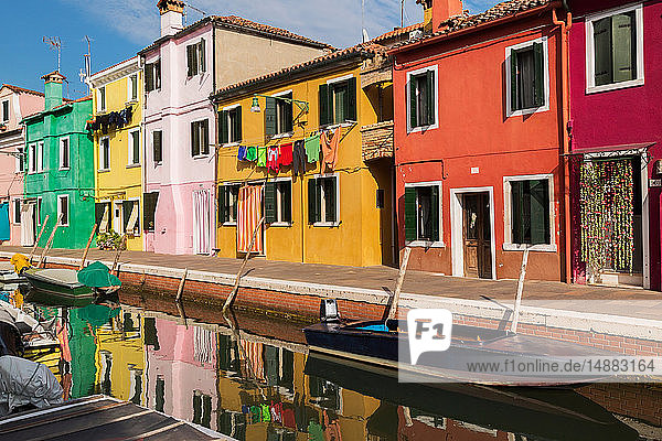 Moored boats on canal lined with pink red orange yellow and green stucco houses decorated with curtains washed clothes on clothesline Burano Island Venetian Lagoon Veneto Italy