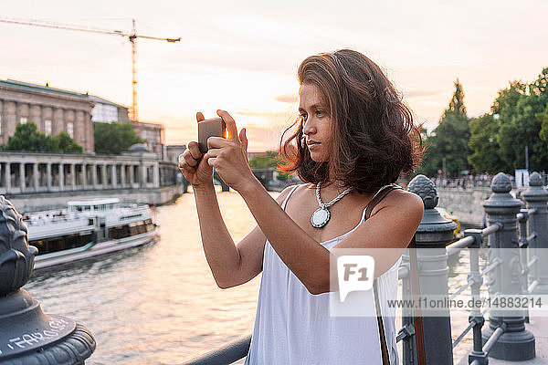 Female student taking photograph with smartphone by river  Berlin  Germany