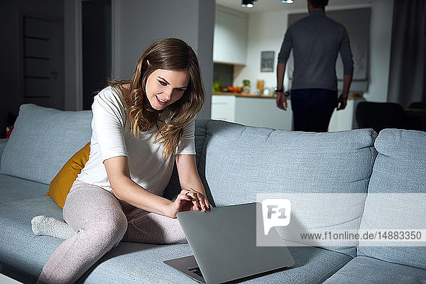 Junge Frau sitzt abends auf dem Sofa und schaut auf den Laptop ihres Freundes