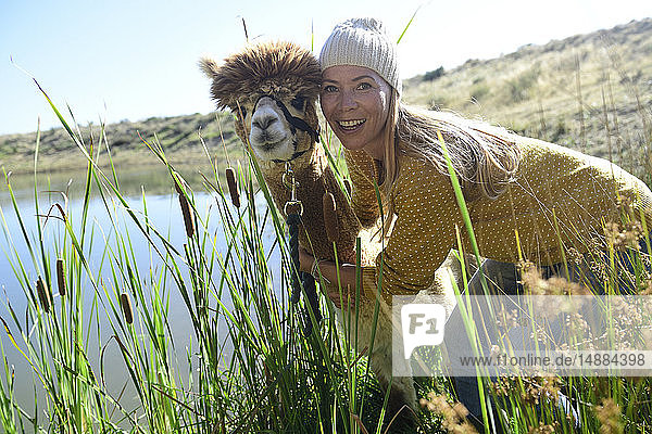 Bildnis einer lächelnden blonden Frau Kopf an Kopf mit Alpaka in der Natur