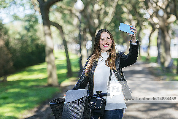 Lächelnde junge Frau mit Fahrrad im Park  die ein Selfie nimmt