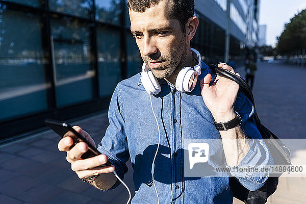 Porträt eines Mannes mit Kopfhörer und Tasche beim Blick auf Handy
