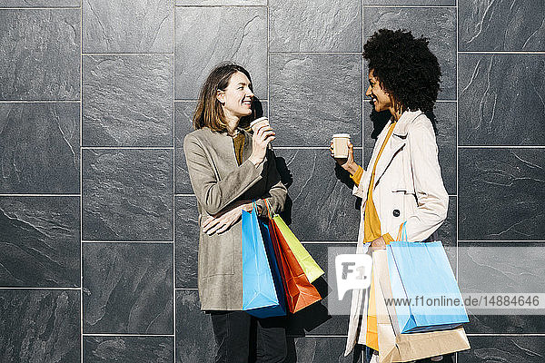 Zwei glückliche Frauen mit Einkaufstaschen und Kaffee zum Mitnehmen stehen an einer Wand und unterhalten sich