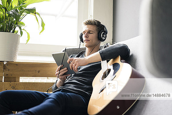 Relaxed young man with tablet and guitar listening to music with headphones