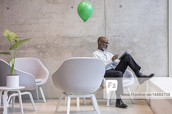 Reifer Geschäftsmann mit grünem Ballon auf Sessel sitzend mit digitalem Tablett