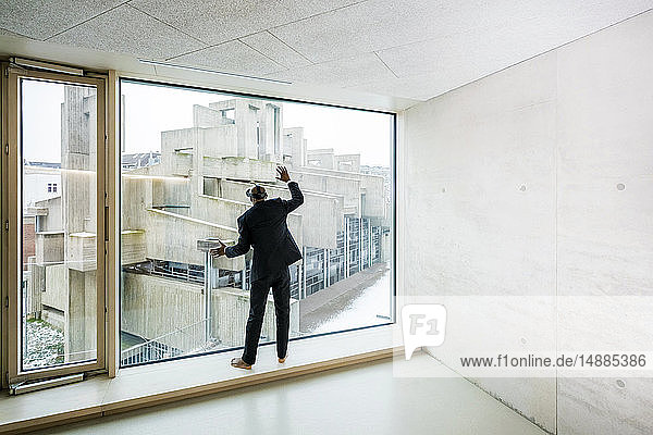 Back view of barefoot businessman with Virtual Reality Glasses standing on window sill looking out of window