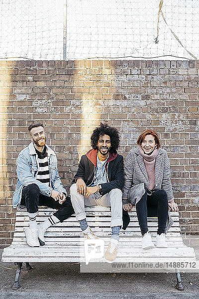 Porträt von drei glücklichen Freunden  die auf einer Bank vor einer Ziegelmauer sitzen