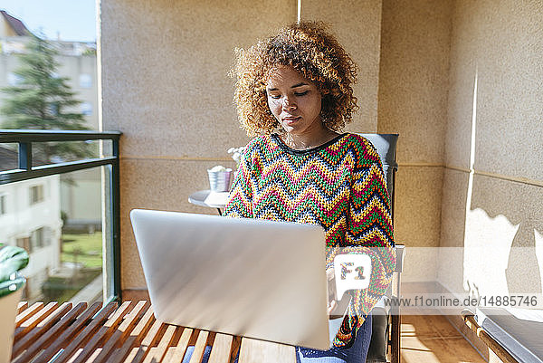 Junge Frau mit lockigem Haar sitzt mit Laptop auf dem Balkon
