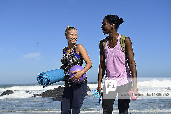 Zwei glückliche Frauen mit Fitnessgeräten am Strand spazieren