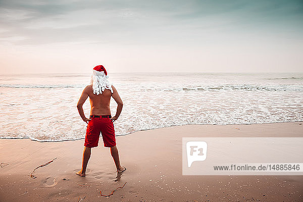 Thailand  Rückenansicht eines als Weihnachtsmann verkleideten Mannes  der bei Sonnenuntergang am Strand steht und auf den Horizont schaut