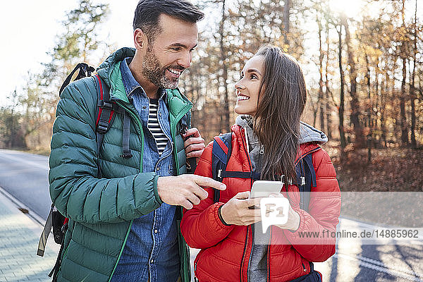 Glückliches Paar überprüft Smartphone während einer Rucksacktour auf einer Straße im Wald
