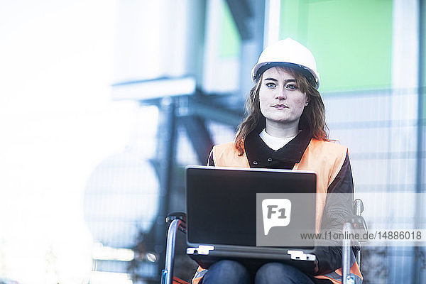 Porträt eines jungen Technikers mit Schutzhelm und Weste im Rollstuhl bei der Arbeit am Laptop im Freien