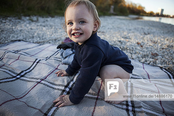 Portrait of smiling toddler girl crouching barefoot on blanket on the beach