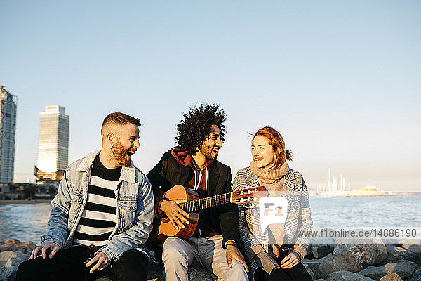 Drei glückliche Freunde mit Gitarre sitzen draußen am Sonnenuntergang an der Küste