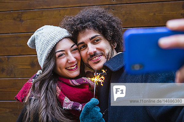 Happy young couple in winterwear holding sparkler and taking a selfie in front of wooden wall