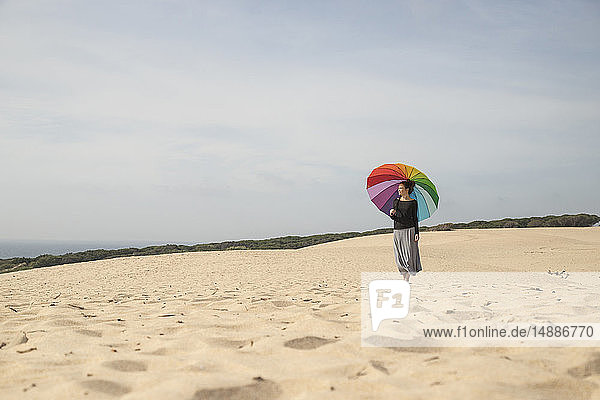 Frau mit buntem Regenschirm steht auf einer Düne und schaut in die Ferne