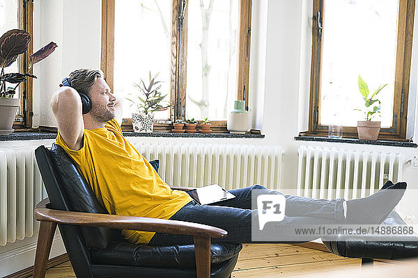 Entspannter Mann in gelbem Hemd mit Kopfhörern sitzt in Lounge Chair in stilvoller Wohnung