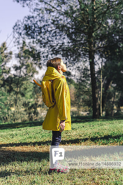 Mädchen in gelbem Regenmantel und gelbem Rucksack steht auf einer Wiese und genießt das Sonnenlicht