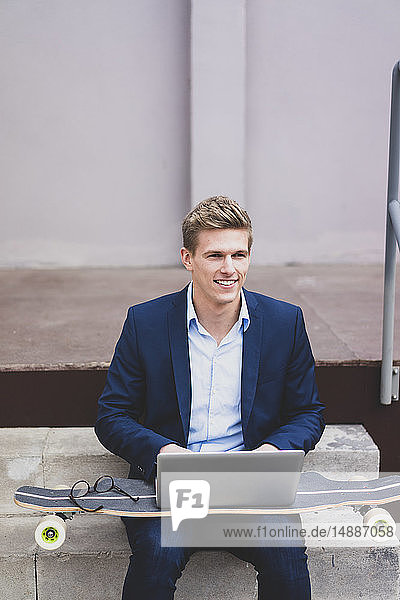 Smiling young businessman with skateboard sitting outdoors on stairs using laptop
