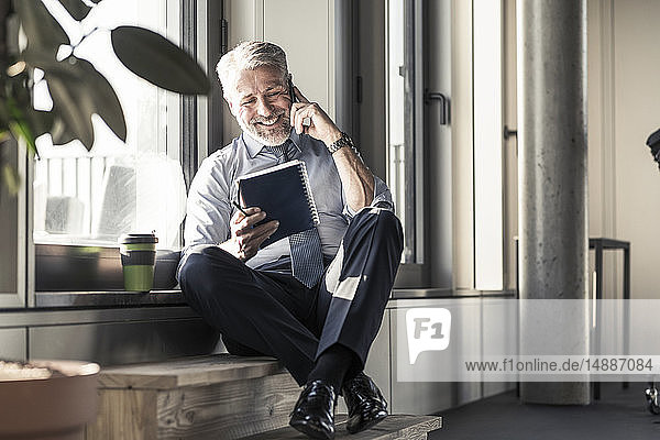 Lächelnder reifer Geschäftsmann mit Notebook  der am Fenster sitzt und mit dem Handy telefoniert