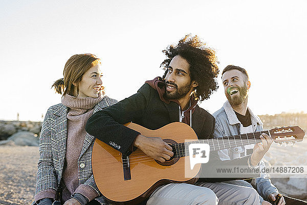 Drei glückliche Freunde mit Gitarre sitzen bei Sonnenuntergang im Freien