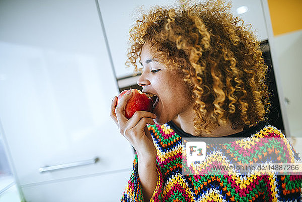 Junge Frau mit lockigem Haar isst einen Apfel in der Küche