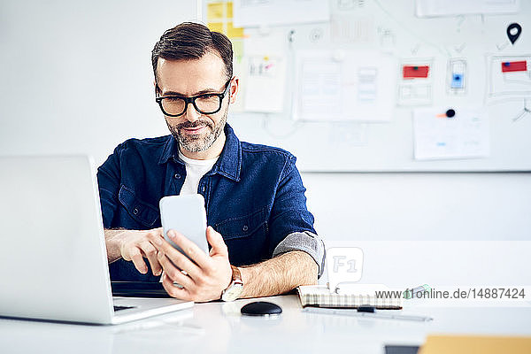 Casual businessman using smartphone and laptop at desk in office