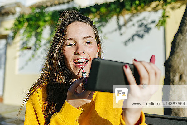 Young woman applying lipstick looking at herself on the cell phone