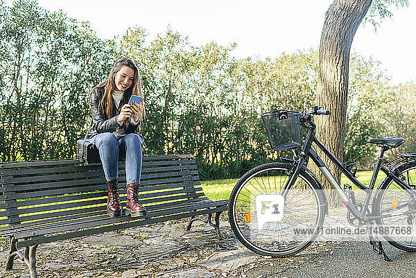 Junge Frau mit Fahrrad sitzt auf einer Parkbank und benutzt ihr Handy