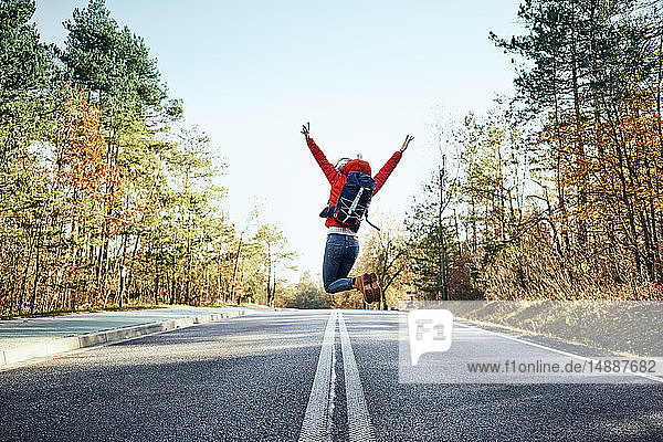 Fröhliche Frau springt während einer Rucksacktour auf eine leere Straße