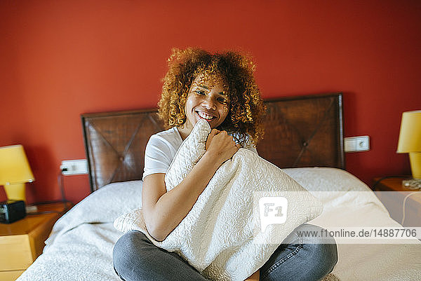 Porträt einer glücklichen jungen Frau mit lockigem Haar  die zu Hause auf dem Bett sitzt und ein Kissen hält