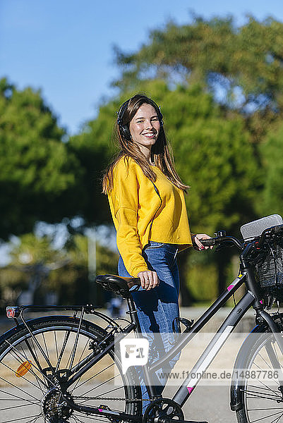 Porträt einer lächelnden jungen Frau mit Fahrrad und Kopfhörer im Park