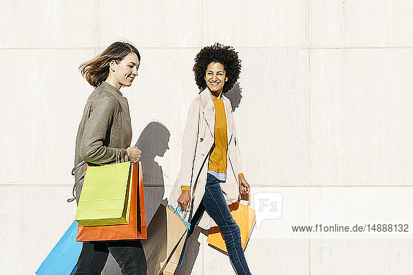 Two happy women with shopping bags walking along a wall