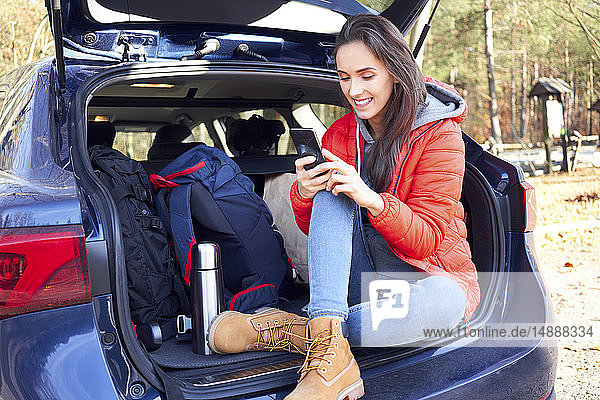 Frau benutzt Smartphone  während sie während einer Autofahrt im Kofferraum sitzt