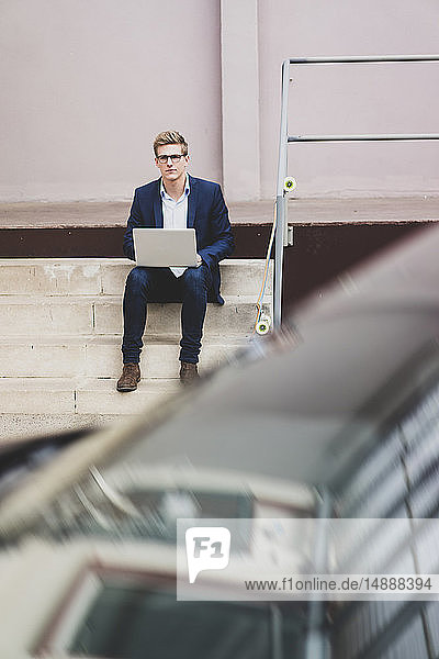 Young businessman with skateboard sitting outdoors on stairs using laptop