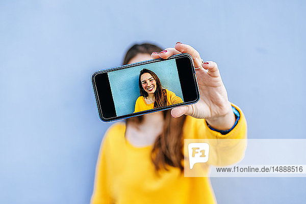Lächelnde junge Frau  die an einer blauen Wand ein Selfie macht