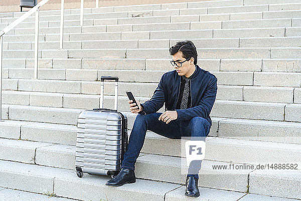 Modischer junger Geschäftsmann mit rollendem Koffer sitzt auf der Treppe und schaut auf sein Handy