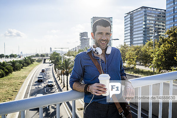 Porträt eines lächelnden Mannes mit Kaffee zum Mitnehmen und Kopfhörern  der sich an das Brückengeländer lehnt
