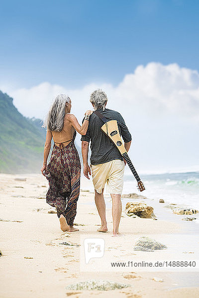 Rückenansicht eines älteren Hippie-Paares mit Gitarre  das Seite an Seite am Strand spazieren geht