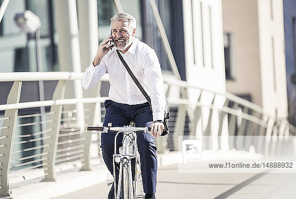 Glücklicher reifer Geschäftsmann  der auf einer Brücke in der Stadt mit dem Handy telefoniert und Fahrrad fährt