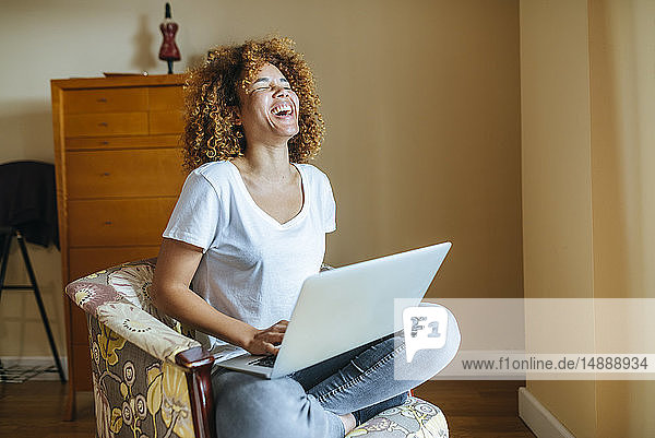 Glückliche junge Frau mit lockigem Haar sitzt zu Hause im Sessel und benutzt ihren Laptop