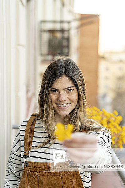 Porträt einer lächelnden jungen Frau auf dem Balkon  die eine gelbe Blüte schenkt