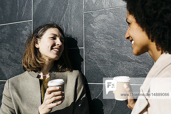 Zwei glückliche Frauen mit Kaffee zum Mitnehmen sprechen gegen eine Wand