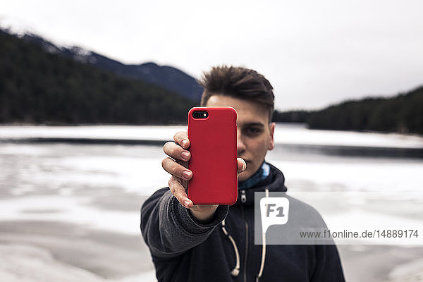 Junger Mann hält im Winter rotes Handy an einem See