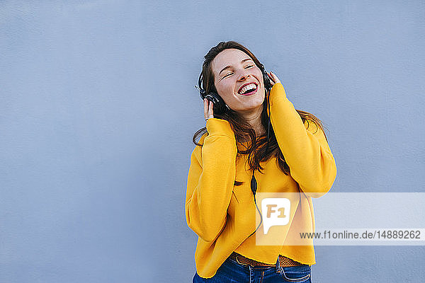 Glückliche junge Frau hört Musik mit Kopfhörern an blauer Wand