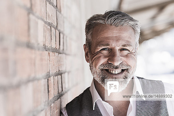 Porträt eines lächelnden reifen Geschäftsmannes  der sich an eine Ziegelwand lehnt