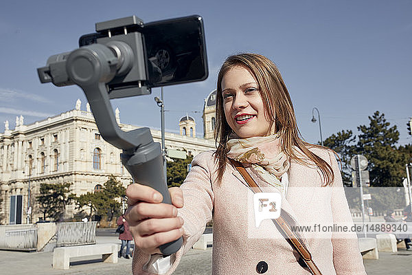 Österreich  Wien  Porträt einer lächelnden jungen Frau  die einen Selfie-Stick zum Fotografieren mit einem Smartphone verwendet
