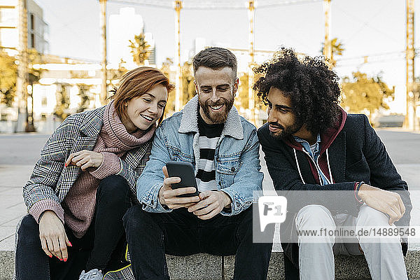 Drei glückliche Freunde sitzen im Freien und schauen auf ihr Handy