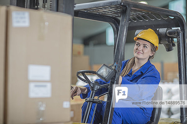Lächelnde Frau mit Schutzhelm fährt Gabelstapler in der Fabrik